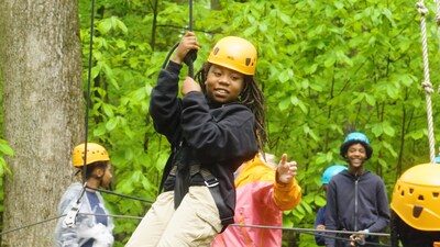 In the woods, surrounded by trees, a young black female Outward Bound Student wearing a climbing helmet is smiling while being lowered by her peers from a ropes element at the Outward Bound campus.