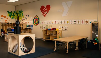 An indoor classroom for children. The room has a play area with a wooden climbing structure, a long table with chairs, and a wall decorated with a large red heart logo, a dartboard, and alphabet cards.