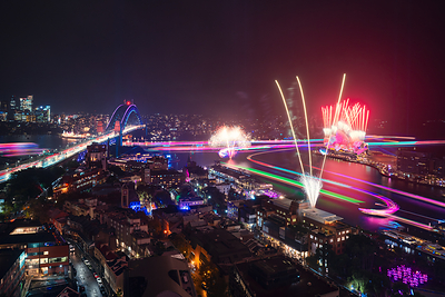 A panoramic night view of Sydney showcasing Vivid Sydney 2026, with fireworks illuminating the sky above the Sydney Opera House and Harbour Bridge